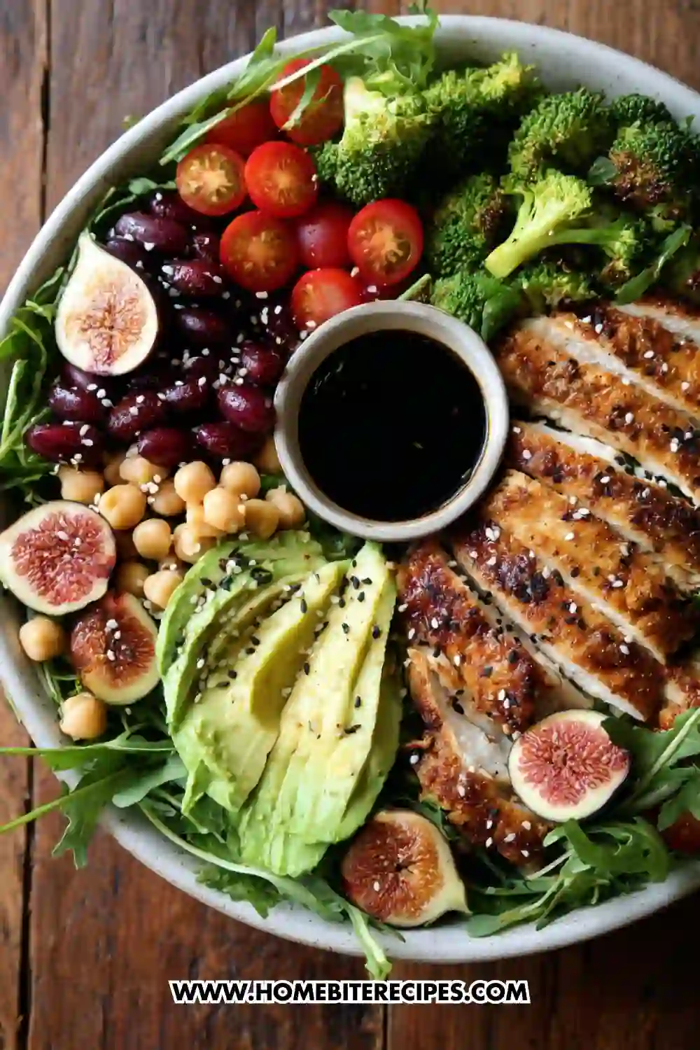 A colorful healthy chicken power bowl filled with sliced grilled chicken, avocado, figs, cherry tomatoes, chickpeas, kidney beans, broccoli, arugula, and a small bowl of balsamic dressing in the center.