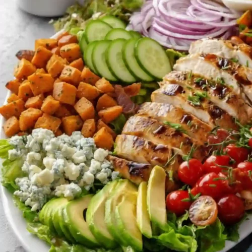 A large white platter filled with a colorful and hearty chicken salad. Ingredients include sliced grilled chicken breast, diced roasted sweet potatoes, sliced avocado, cherry tomatoes, crumbled blue cheese, sliced cucumber, red onion, and hard-boiled egg slices, all arranged over a bed of mixed greens. A small pitcher of dressing is in the background.