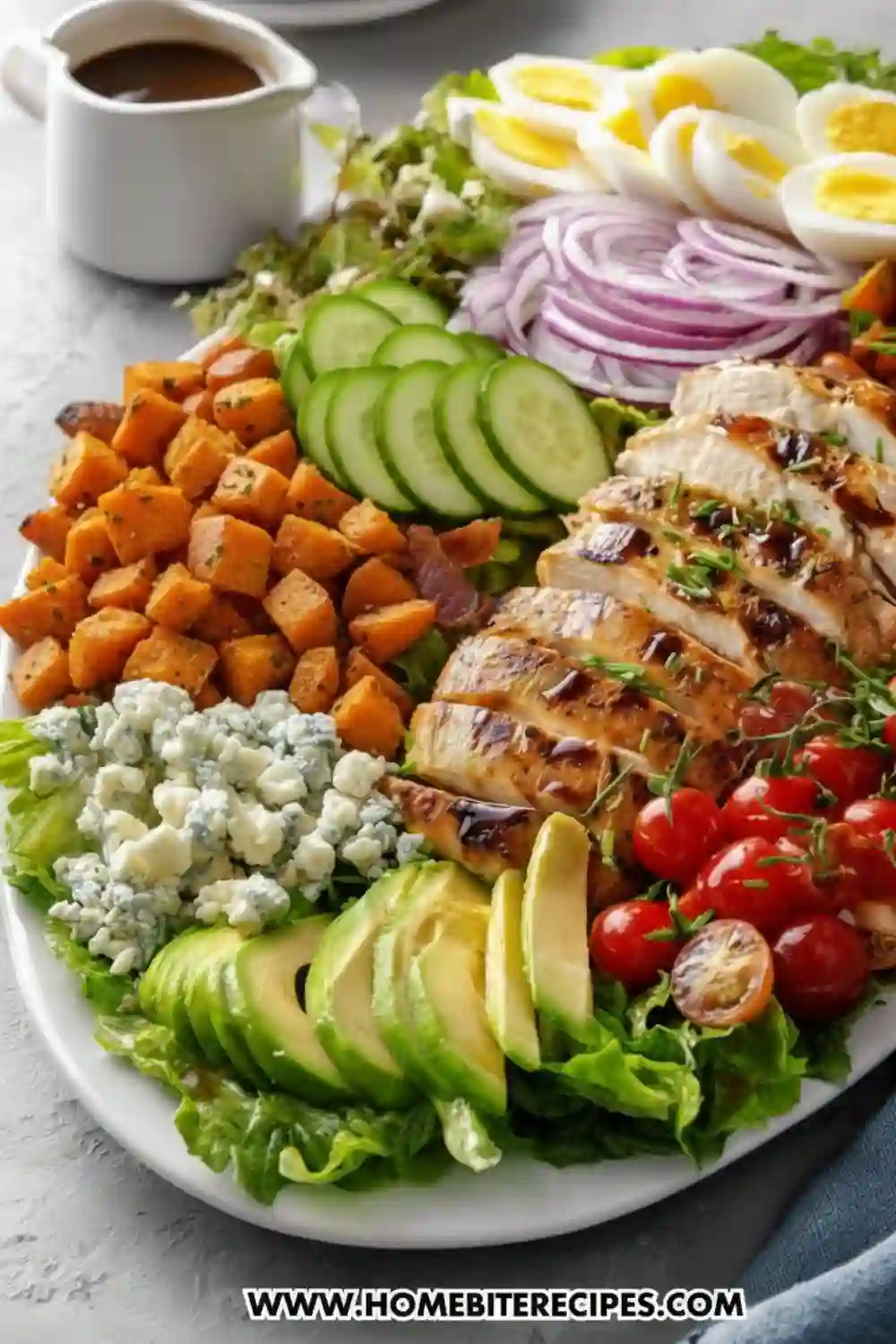 A large white platter filled with a colorful and hearty chicken salad. Ingredients include sliced grilled chicken breast, diced roasted sweet potatoes, sliced avocado, cherry tomatoes, crumbled blue cheese, sliced cucumber, red onion, and hard-boiled egg slices, all arranged over a bed of mixed greens. A small pitcher of dressing is in the background.