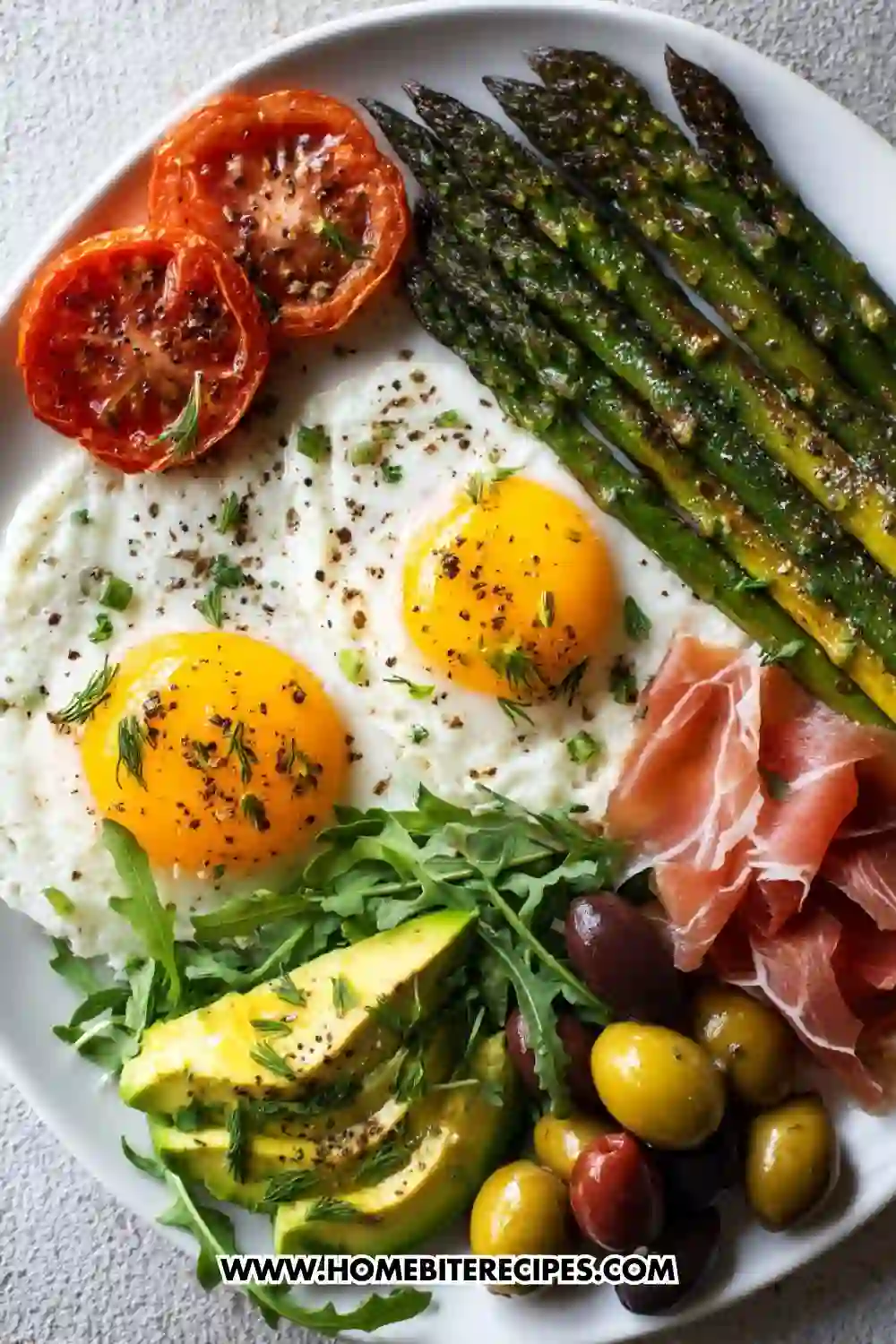 Colorful Mediterranean breakfast plate with sunny-side eggs, roasted asparagus, blistered tomatoes, avocado slices, olives, arugula, and prosciutto on a white dish
