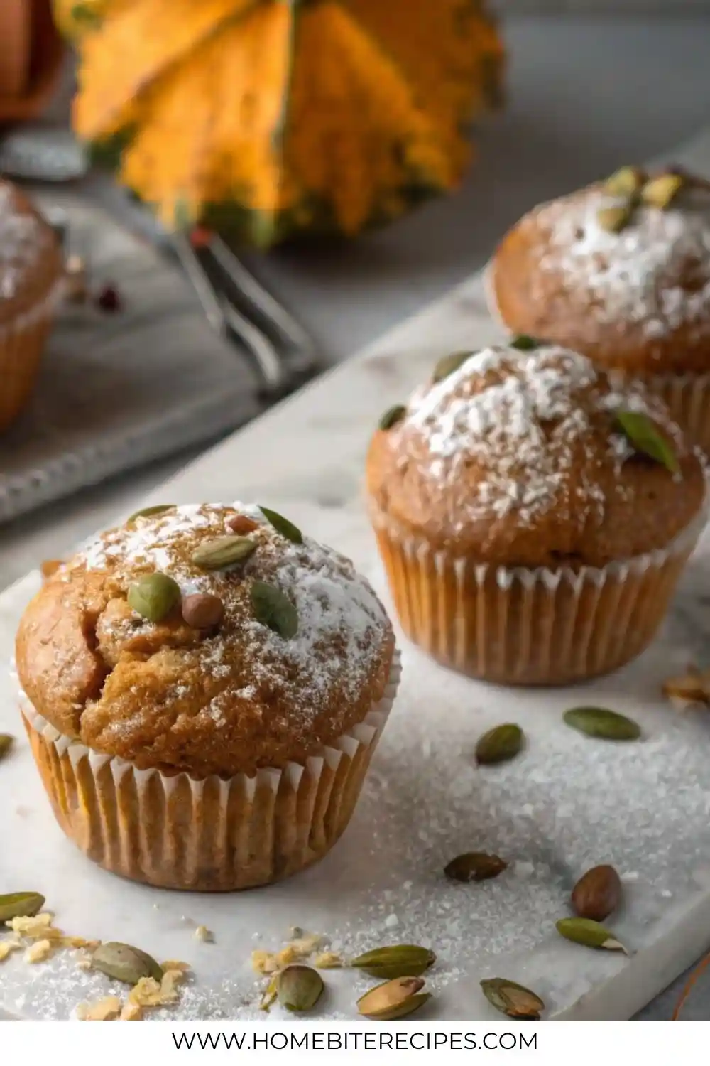 Close-up of freshly baked pumpkin muffins topped with powdered sugar and pumpkin seeds, arranged on a marble board with scattered seeds and fall décor in the background.