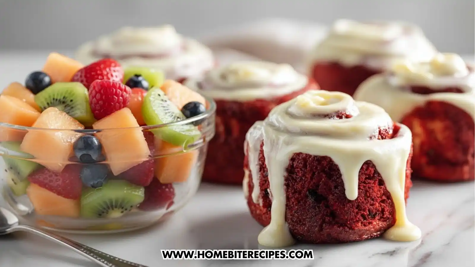 Close-up of Red Velvet Cinnamon Rolls topped with creamy white frosting, showcasing their rich red color and gooey texture served with a simple fruit salad with melon, kiwi, and berries tossed in a touch of honey and lemon juice.
