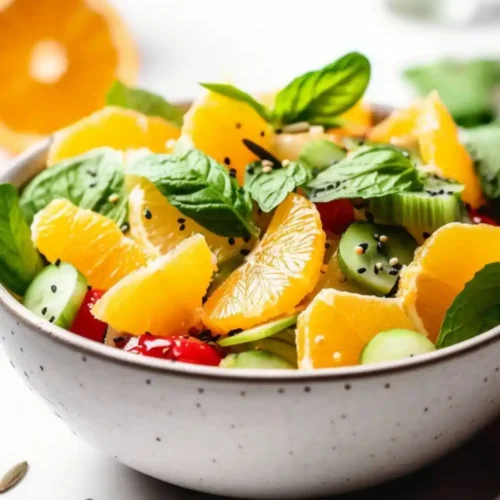 A vibrant bowl of citrus cucumber salad featuring juicy orange slices, fresh mint leaves, cherry tomatoes, and sliced cucumbers, garnished with sesame seeds and served in a white ceramic bowl.