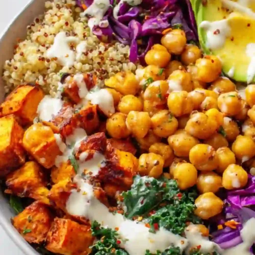 A vibrant vegan Buddha bowl filled with roasted sweet potatoes, chickpeas, quinoa, avocado, kale, and purple cabbage drizzled with creamy tahini dressing.