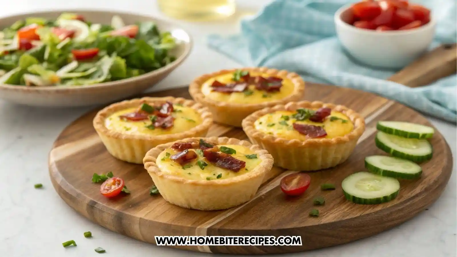 A close-up of four savory mini quiche appetizers with golden flaky crust, creamy egg filling, crispy bacon, and fresh chives on top, arranged on a wooden board with slices of cucumber