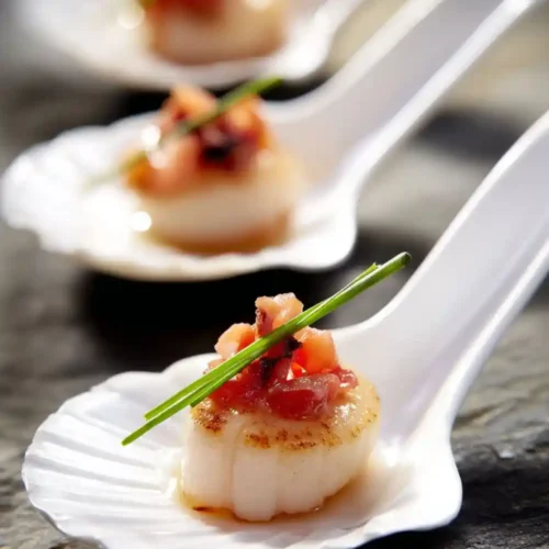 Close-up of seared scallops topped with crispy bacon and chive garnish served on white appetizer spoons, perfect elegant seafood starter.
