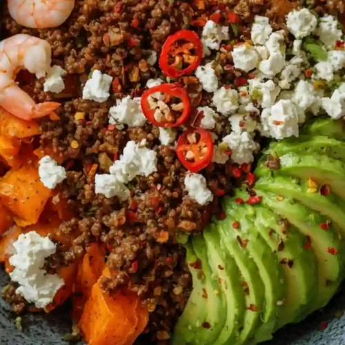 Close-up of a colorful power bowl filled with spicy ground beef, cooked shrimp, roasted sweet potatoes, sliced avocado, feta cheese, and red chili slices in a rustic ceramic dish.