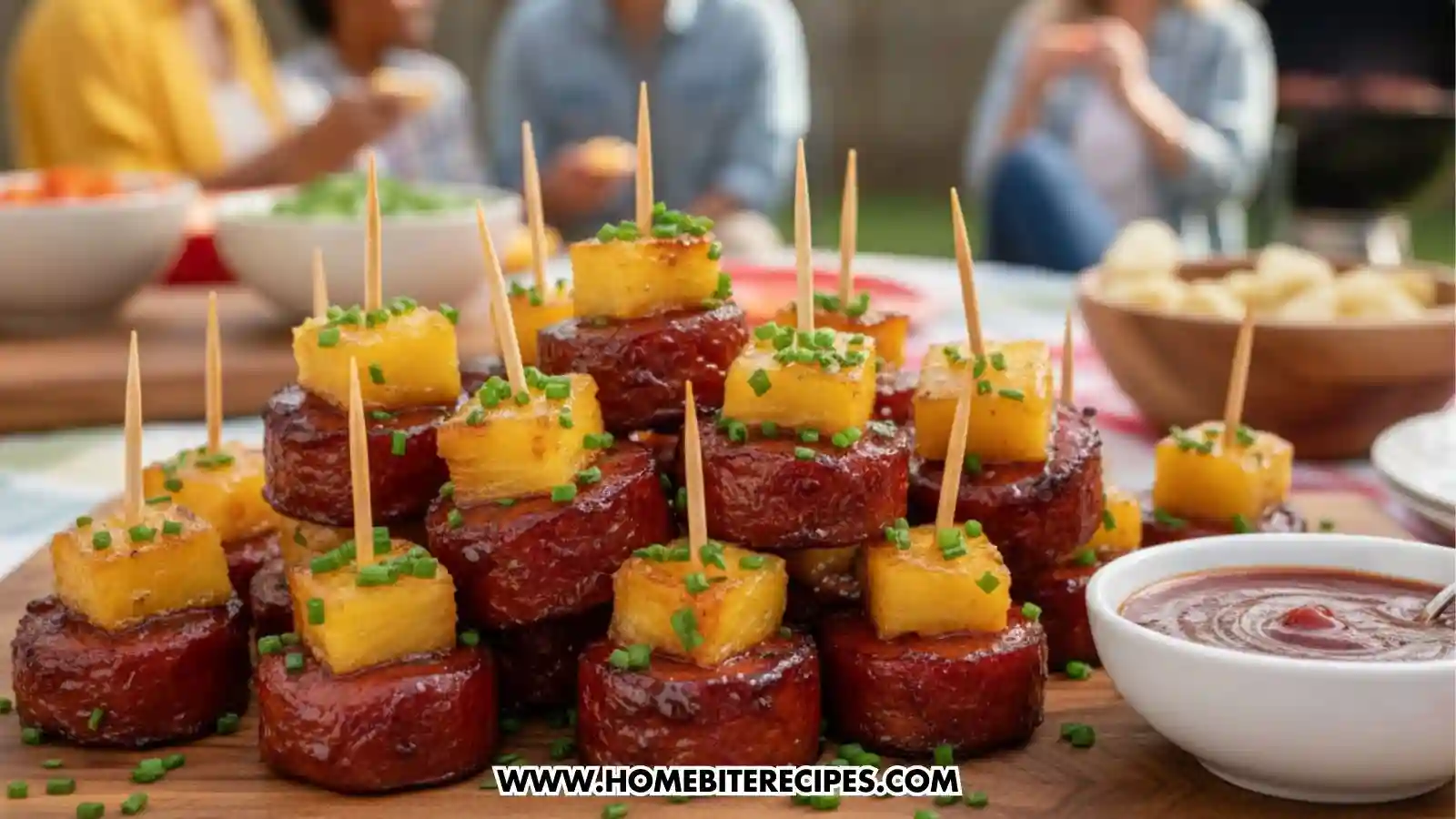 A large stack of sticky glazed smoked sausage bites served on a wooden board at an outdoor BBQ. Each round sausage slice is topped with a golden cube of grilled pineapple, secured with a toothpick, and garnished with chopped green chives. A small white bowl of BBQ sauce sits to the right, and blurred figures of people eating at a picnic table are visible in the background.