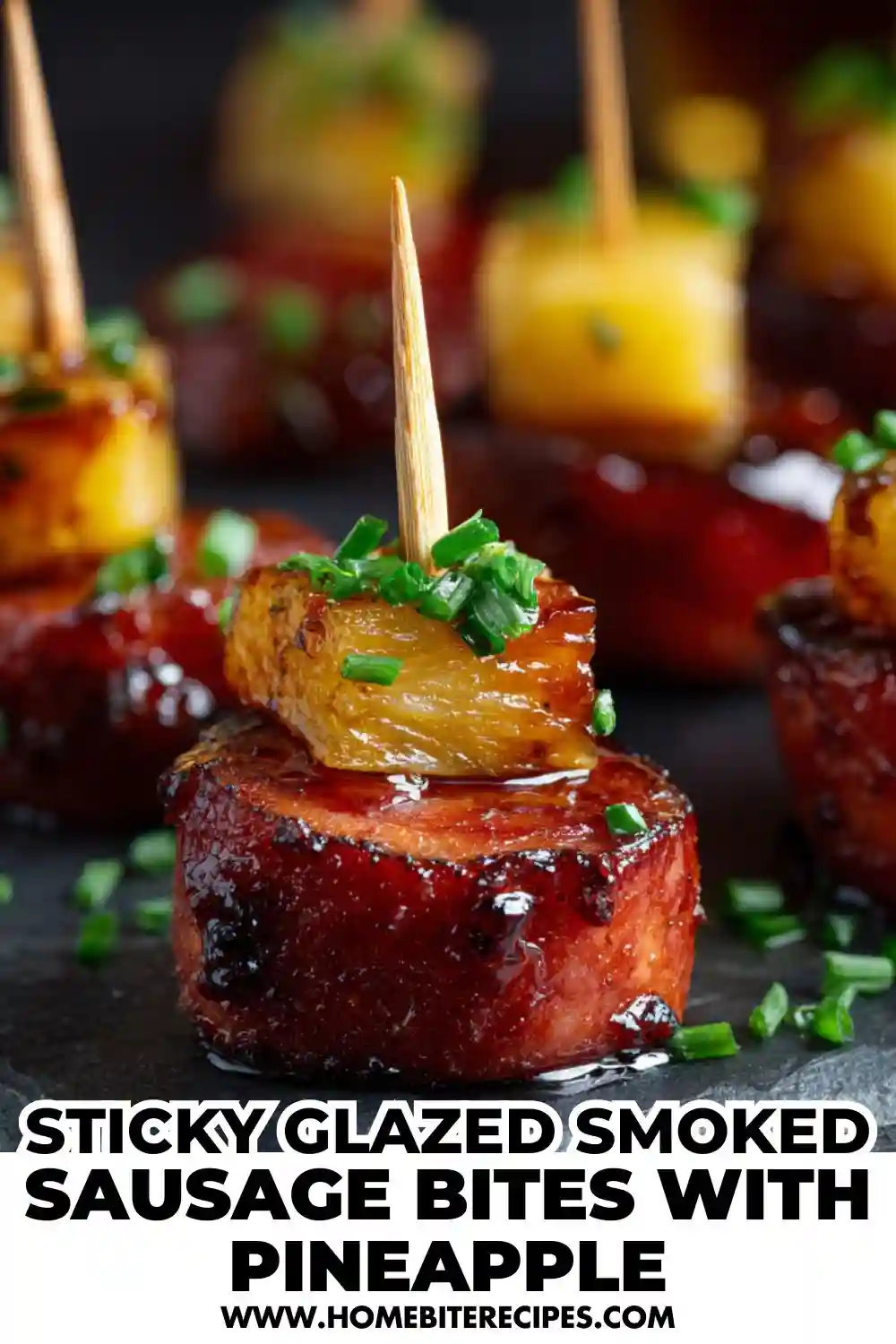 Close-up of sticky glazed smoked sausage bites topped with caramelized pineapple chunks and chopped chives, served as a party appetizer.