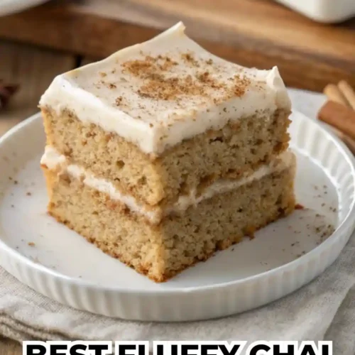A square slice of two-layer chai spiced cake with thick white frosting on a white plate, garnished with cinnamon sticks and star anise on a wooden table.