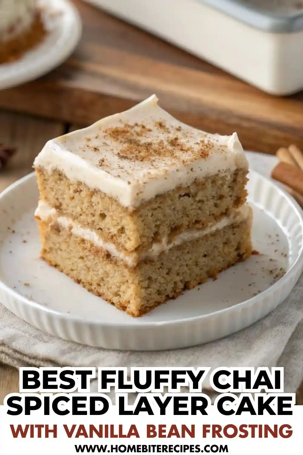 A square slice of two-layer chai spiced cake with thick white frosting on a white plate, garnished with cinnamon sticks and star anise on a wooden table.