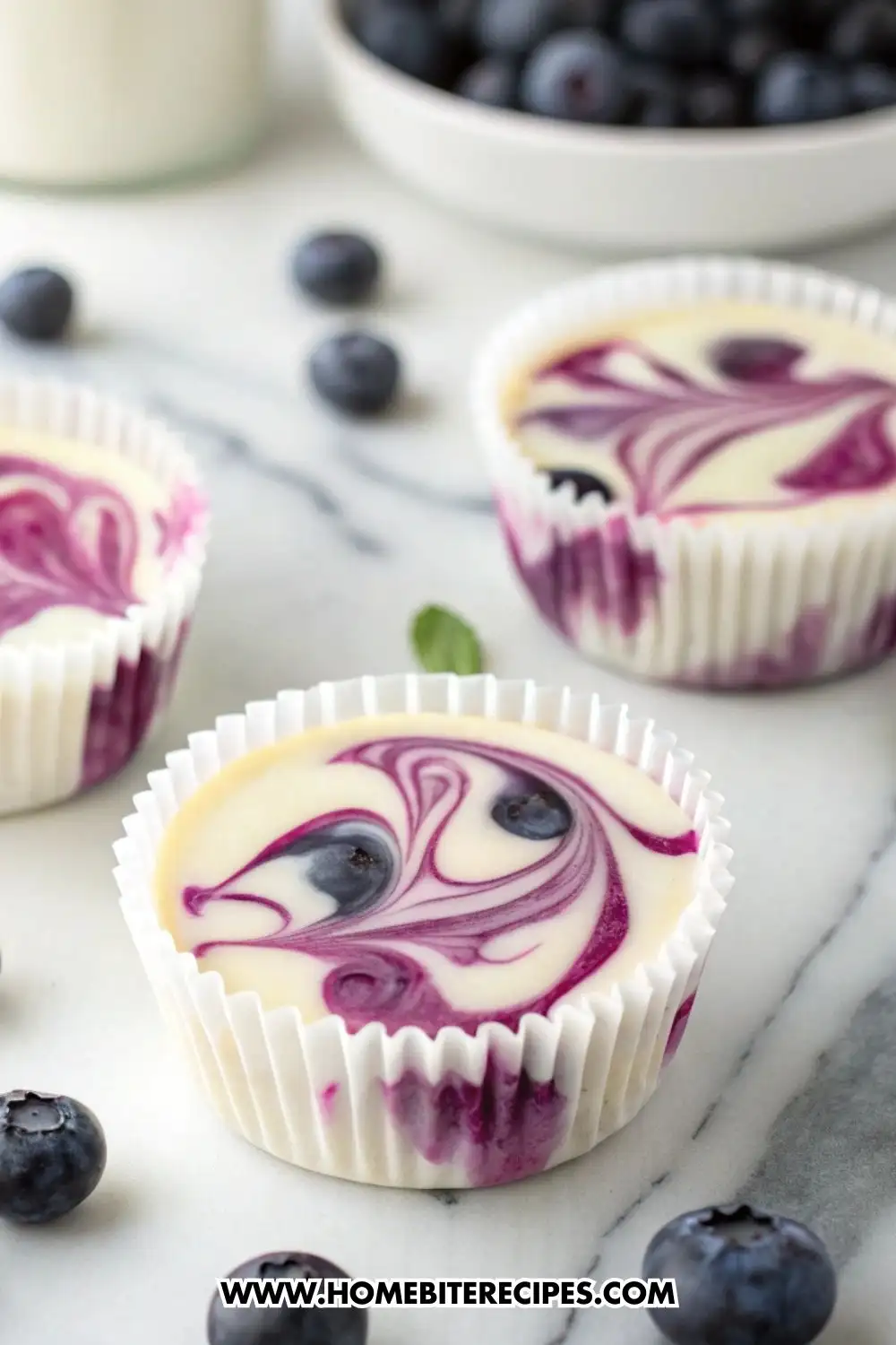 Mini blueberry swirl cheesecake cups in white muffin liners on a marble surface, topped with purple blueberry puree swirls and fresh whole blueberries around them.