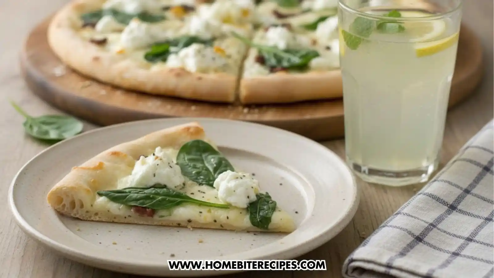A close-up of a single slice of Cheesy White Garlic Pizza with Spinach on a cream-colored plate, topped with fresh spinach leaves and creamy ricotta cheese. In the background, a whole pizza sits on a wooden board next to a tall glass of iced lemonade garnished with mint and a lemon slice.