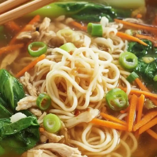 A close-up bowl of Chicken Noodle Soup with Ginger Scallion featuring ramen noodles, shredded chicken, sliced carrots, and fresh bok choy.
