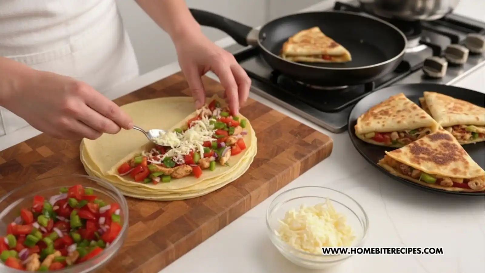 A person’s hands assembling quesadillas on a wooden cutting board. They are using a spoon to add shredded cheese over a filling of diced chicken, red and green bell peppers, and onions on a round tortilla. In the background, a quesadilla cooks in a skillet on a stovetop, and a plate of finished golden-brown wedges sits to the right.