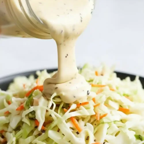 Creamy Classic Homemade Coleslaw Dressing being poured from a glass jar onto a fresh bowl of shredded cabbage and carrots.