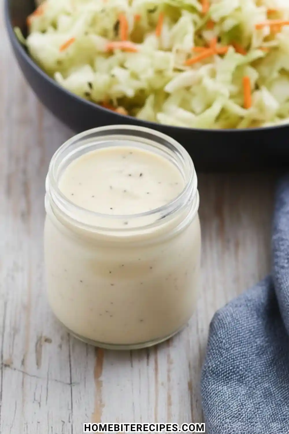 A glass jar filled with creamy Classic Homemade Coleslaw Dressing speckled with black pepper, sitting next to a bowl of fresh cabbage.