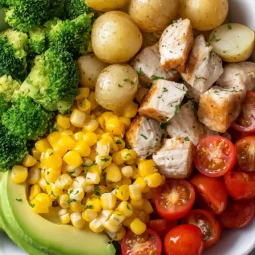 Clean eating chicken bowl with grilled chicken, baby potatoes, broccoli, corn, avocado slices, and cherry tomatoes.