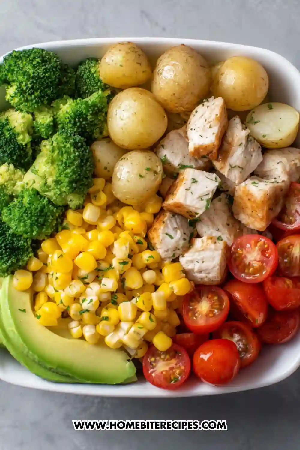 Clean eating chicken bowl with grilled chicken, baby potatoes, broccoli, corn, avocado slices, and cherry tomatoes.