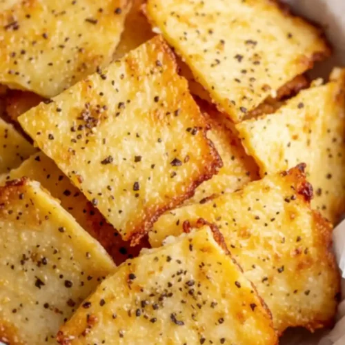 A close-up of crispy, golden-brown Cottage Cheese Chips seasoned with cracked black pepper in a wooden bowl.