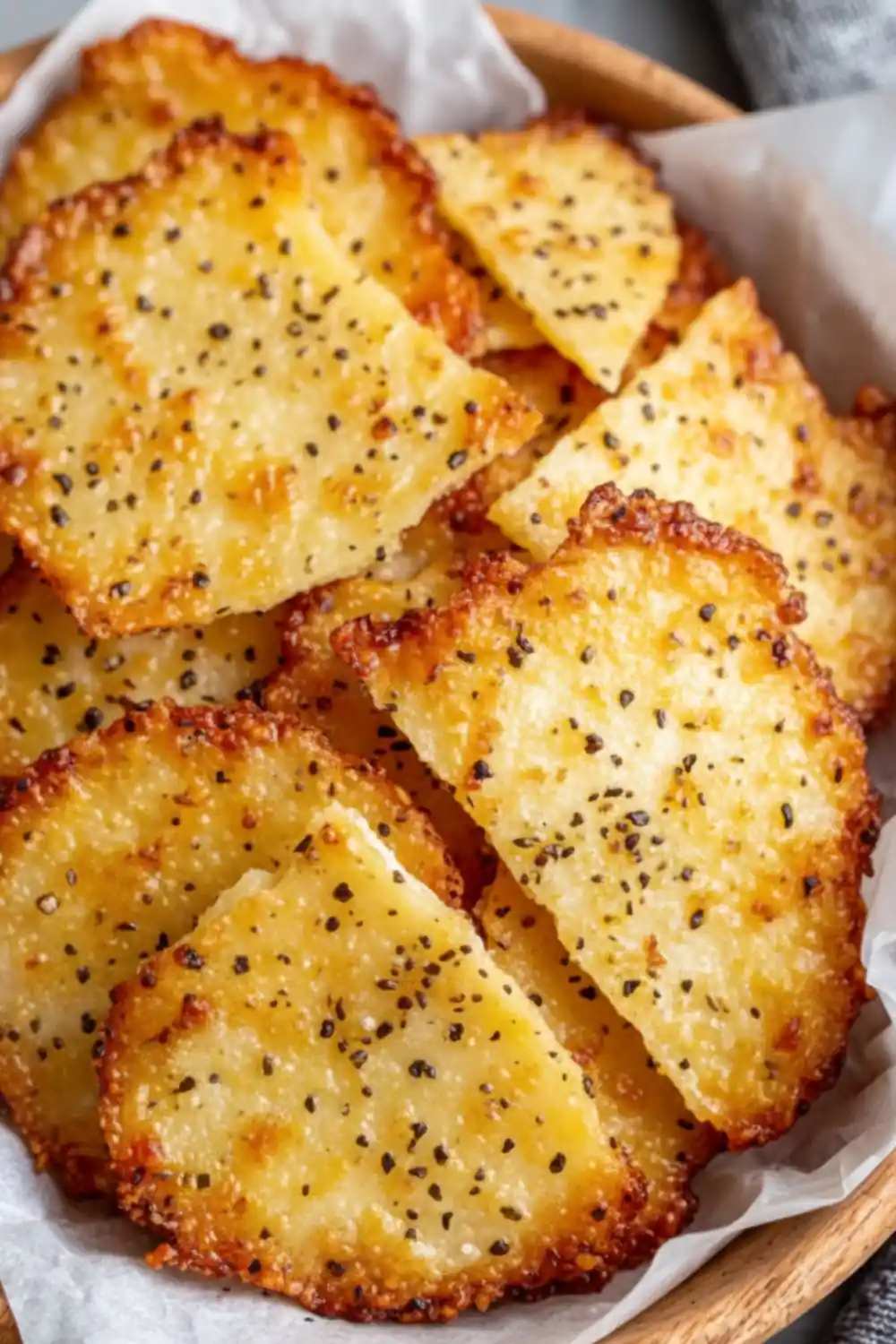 A close-up of crispy, golden-brown Cottage Cheese Chips seasoned with cracked black pepper in a wooden bowl.