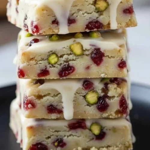 A tall stack of four square Cranberry Pistachio Shortbread Cookies featuring a white chocolate glaze, dried cranberries, and chopped green pistachios.