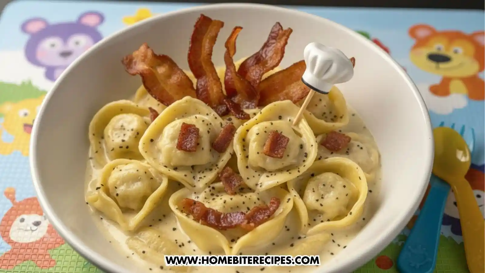 A playful kid-friendly presentation of tortellini Alfredo in a white bowl. The tortellini are arranged like faces with small bacon bits for eyes and smiles. Strips of crispy bacon are placed at the top to look like hair, and a small decorative chef's hat toothpick is inserted into one pasta piece. The bowl sits on a colorful cartoon-themed placemat.