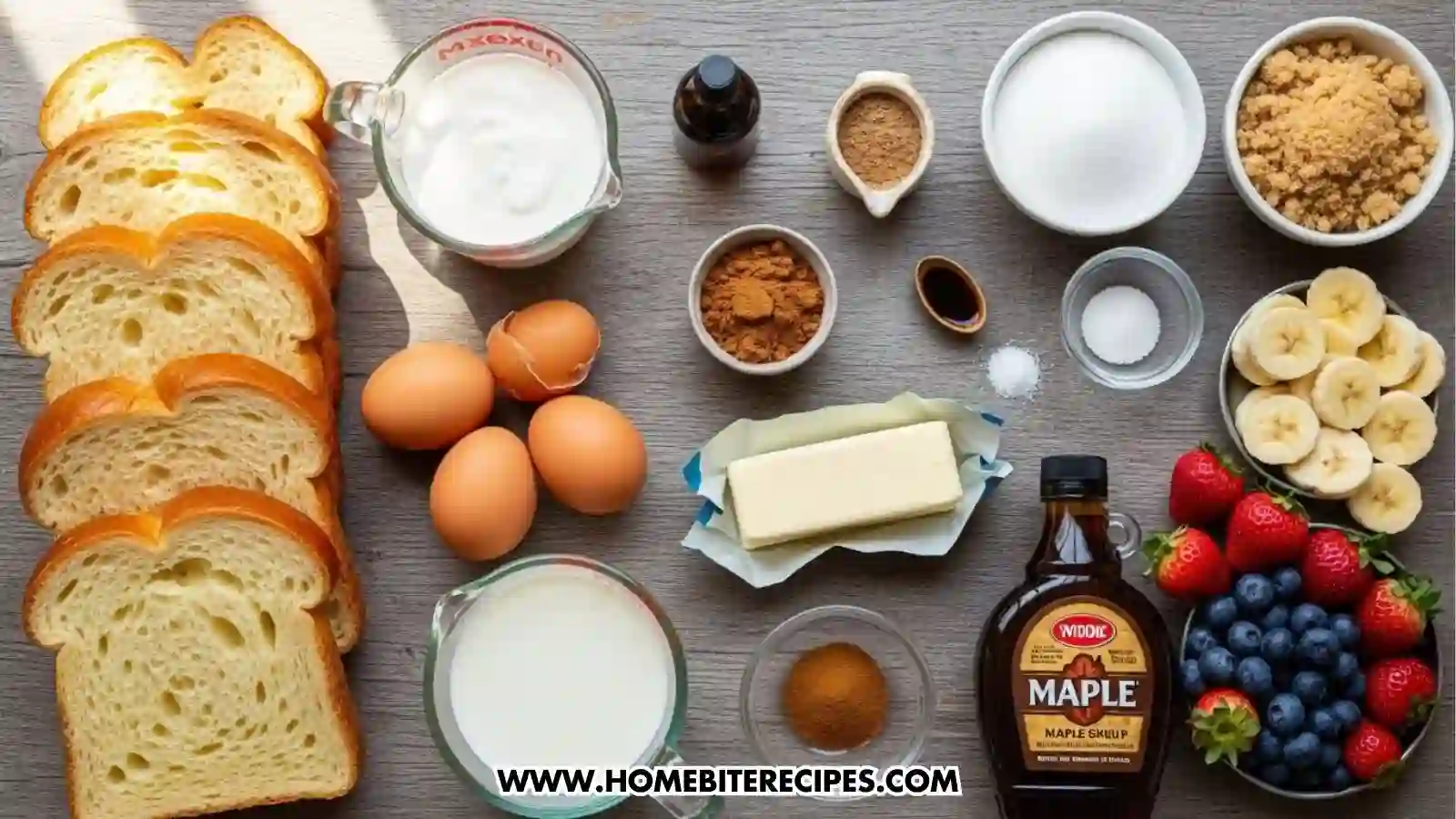 A top-down overhead view of various ingredients on a grey wooden surface. Included are thick slices of brioche bread, four brown eggs, a carton of heavy cream, a bowl of milk, butter, vanilla extract, cinnamon, nutmeg, brown sugar, white sugar, maple syrup, and bowls of fresh strawberries, blueberries, and sliced bananas.
