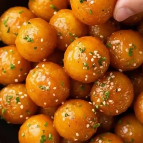 A close-up view of golden crispy potato balls glazed with garlic butter, sprinkled with sesame seeds and chopped herbs, with one ball being picked up by hand.