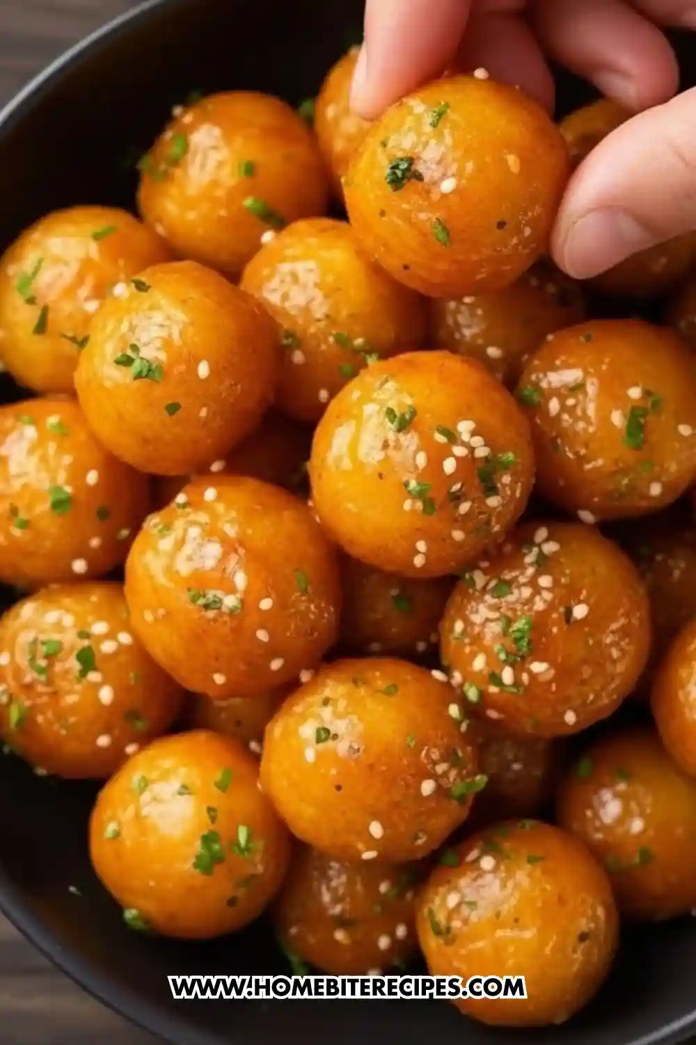 A close-up view of golden crispy potato balls glazed with garlic butter, sprinkled with sesame seeds and chopped herbs, with one ball being picked up by hand.