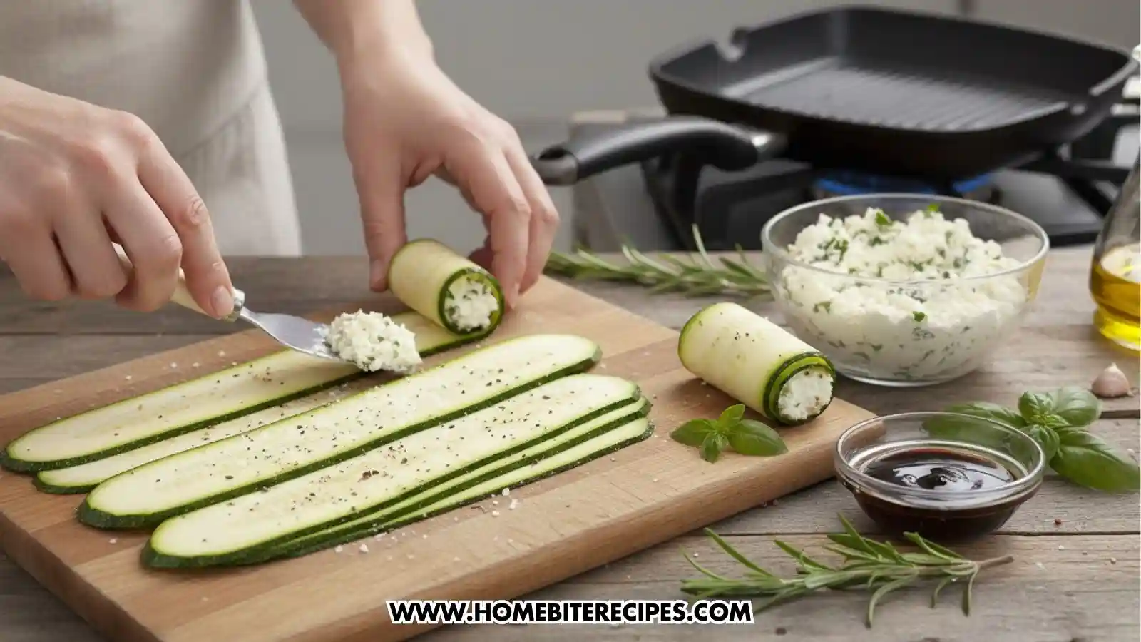 A person’s hands using a small spatula to spread a ricotta cheese mixture onto thin, seasoned slices of zucchini on a wooden cutting board. Several rolls are already formed, with a bowl of herb-flecked cheese, a small dish of balsamic glaze, and a grill pan on a stovetop visible in the background.