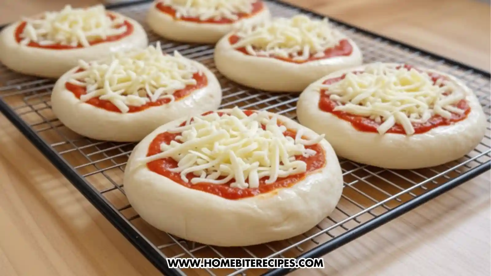 A close-up, slightly angled view of six mini Easy Cheesy Pizza Pockets being prepared on a black wire cooling rack. Each pocket consists of a thick, round base of white dough topped with a circular layer of bright red tomato sauce and a generous pile of shredded mozzarella cheese. The rack sits on a light-colored wooden countertop, showing the pizzas in various stages of focus from front to back.