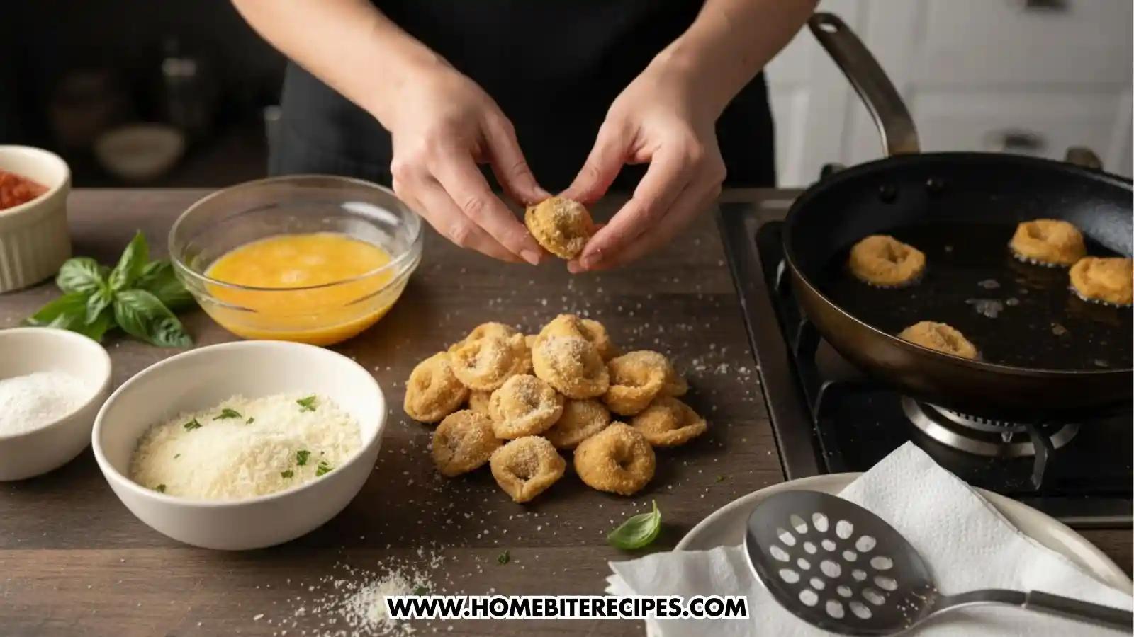 A person preparing fried tortellini on a dark countertop. They are shown coating a piece of pasta in breadcrumbs. The scene includes a bowl of egg wash, a bowl of parmesan breading, a frying pan with tortellini cooking in oil on a stovetop, and a slotted spoon resting on paper towels.