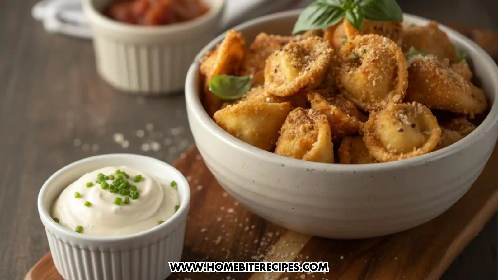 A close-up shot of a white bowl filled with golden-brown, breaded fried tortellini garnished with fresh basil. In the foreground, there is a small white ramekin of creamy white garlic sauce topped with chopped chives. A small dish of red marinara sauce is visible in the background on a wooden serving board.