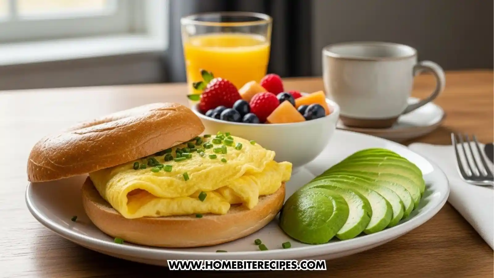 A bright brunch plate featuring a bagel sandwich filled with a fluffy folded egg topped with chopped chives. The plate includes fanned avocado slices and is served with a side bowl of mixed fruit, a glass of orange juice, and a cup of coffee in the background.