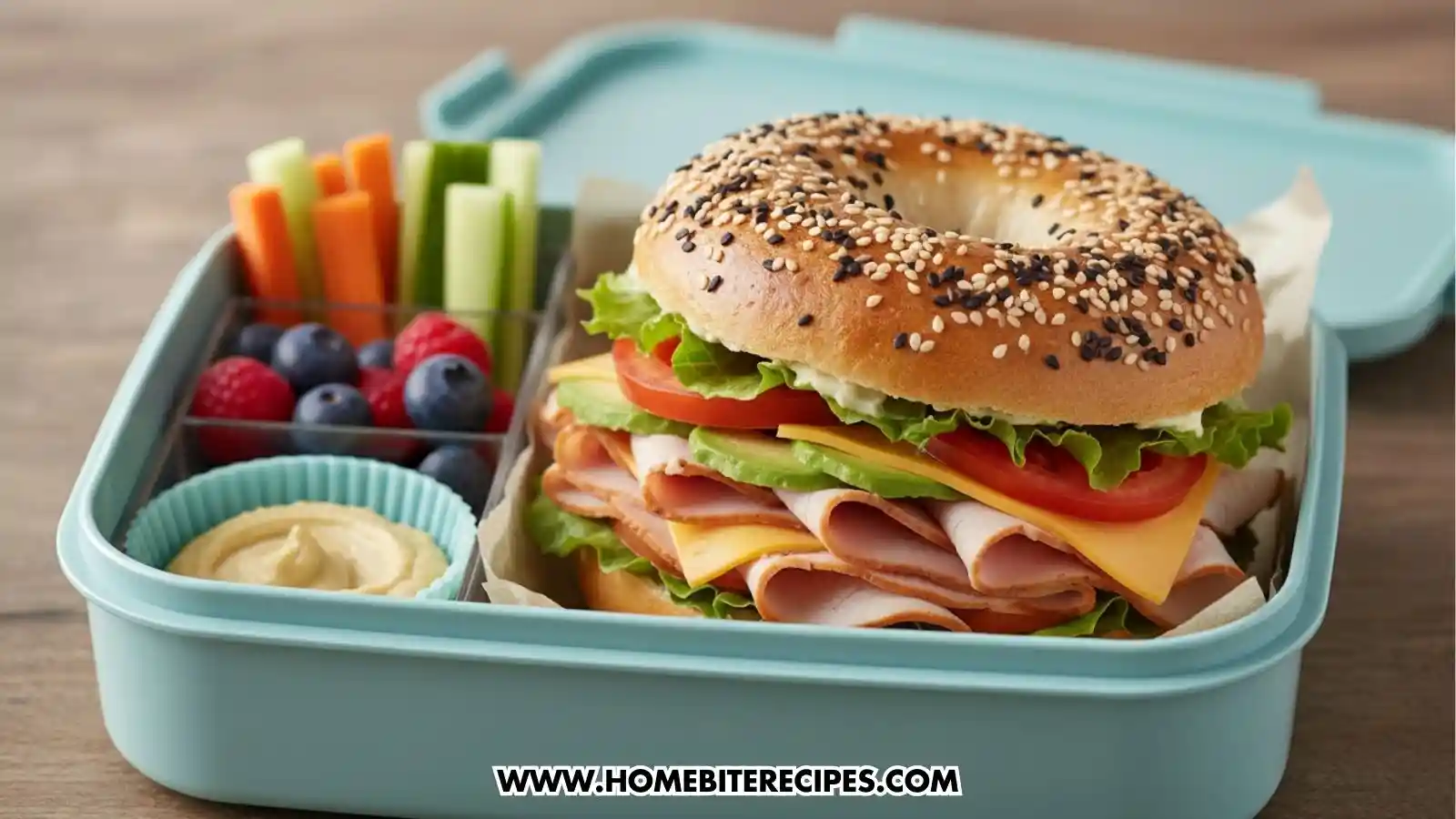 A light blue bento-style lunch box packed with a large bagel sandwich loaded with turkey, cheese, tomato, lettuce, and avocado. The side compartments contain carrot and celery sticks, fresh blueberries and raspberries, and a small cup of hummus.