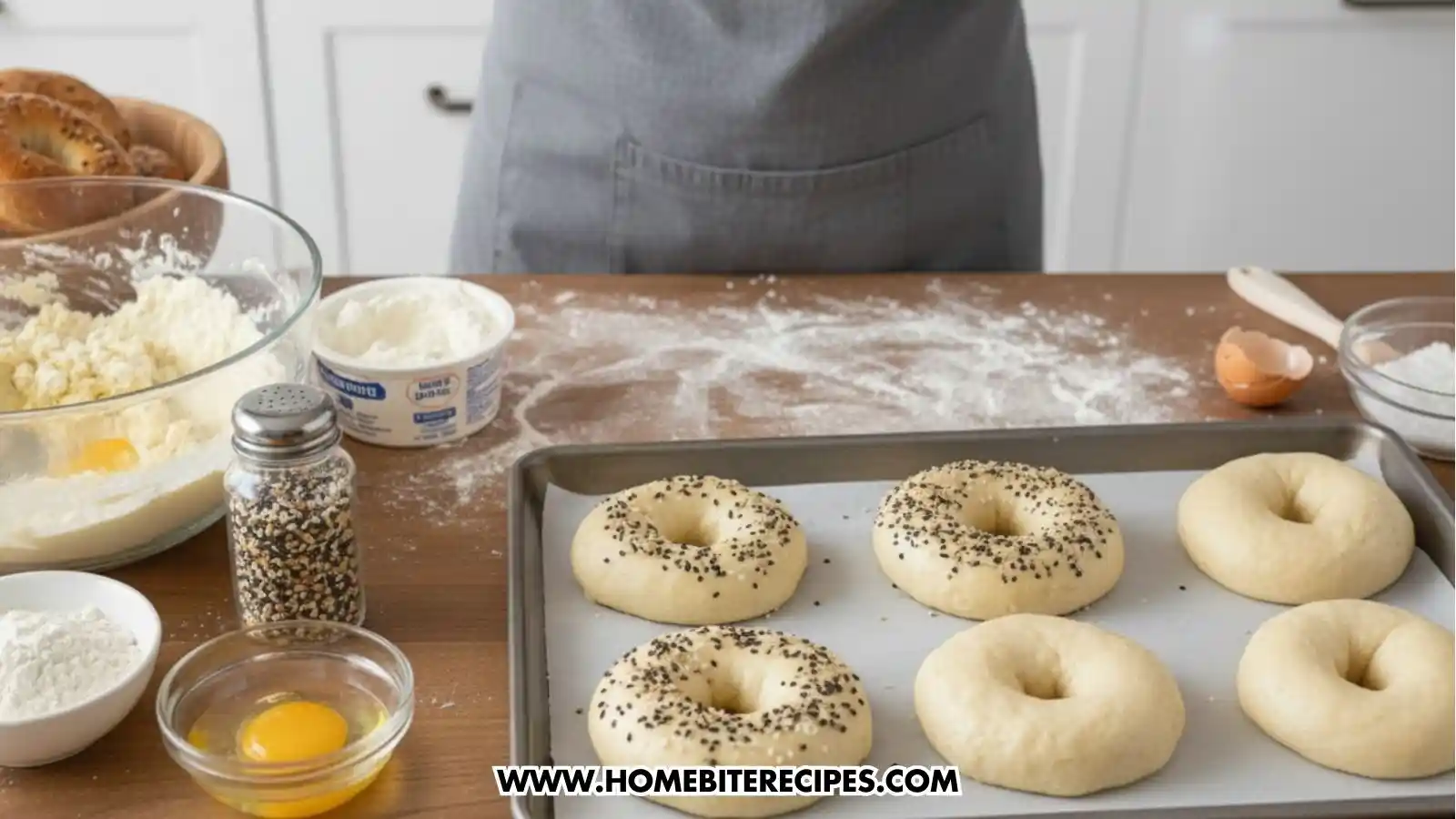 A person in a grey apron preparing bagels on a wooden counter. On a baking sheet, four raw dough bagels are visible—two topped with sesame seeds and two plain. The counter features ingredients including a bowl of almond flour, a container of cottage cheese, an open egg, and a jar of bagel seasoning.