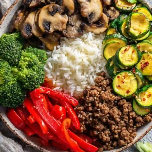 A colorful rice bowl filled with sautéed mushrooms, cooked zucchini, fresh broccoli, red bell peppers, and seasoned ground beef arranged around a mound of white rice.