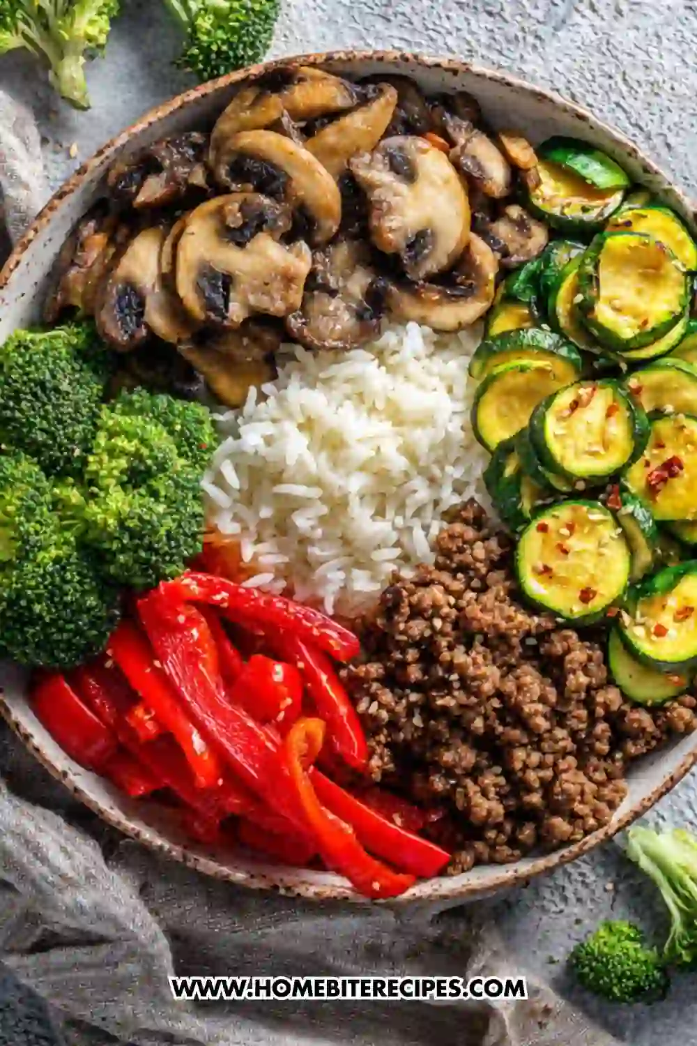 A colorful rice bowl filled with sautéed mushrooms, cooked zucchini, fresh broccoli, red bell peppers, and seasoned ground beef arranged around a mound of white rice.