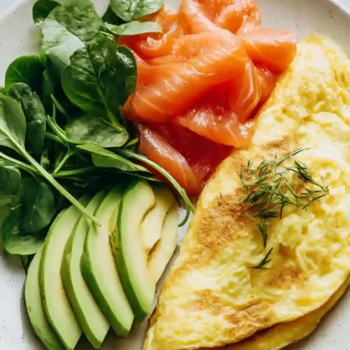Healthy omelet served with smoked salmon, sliced avocado, and fresh spinach on a ceramic plate, topped with dill and prepared as a low-carb breakfast.