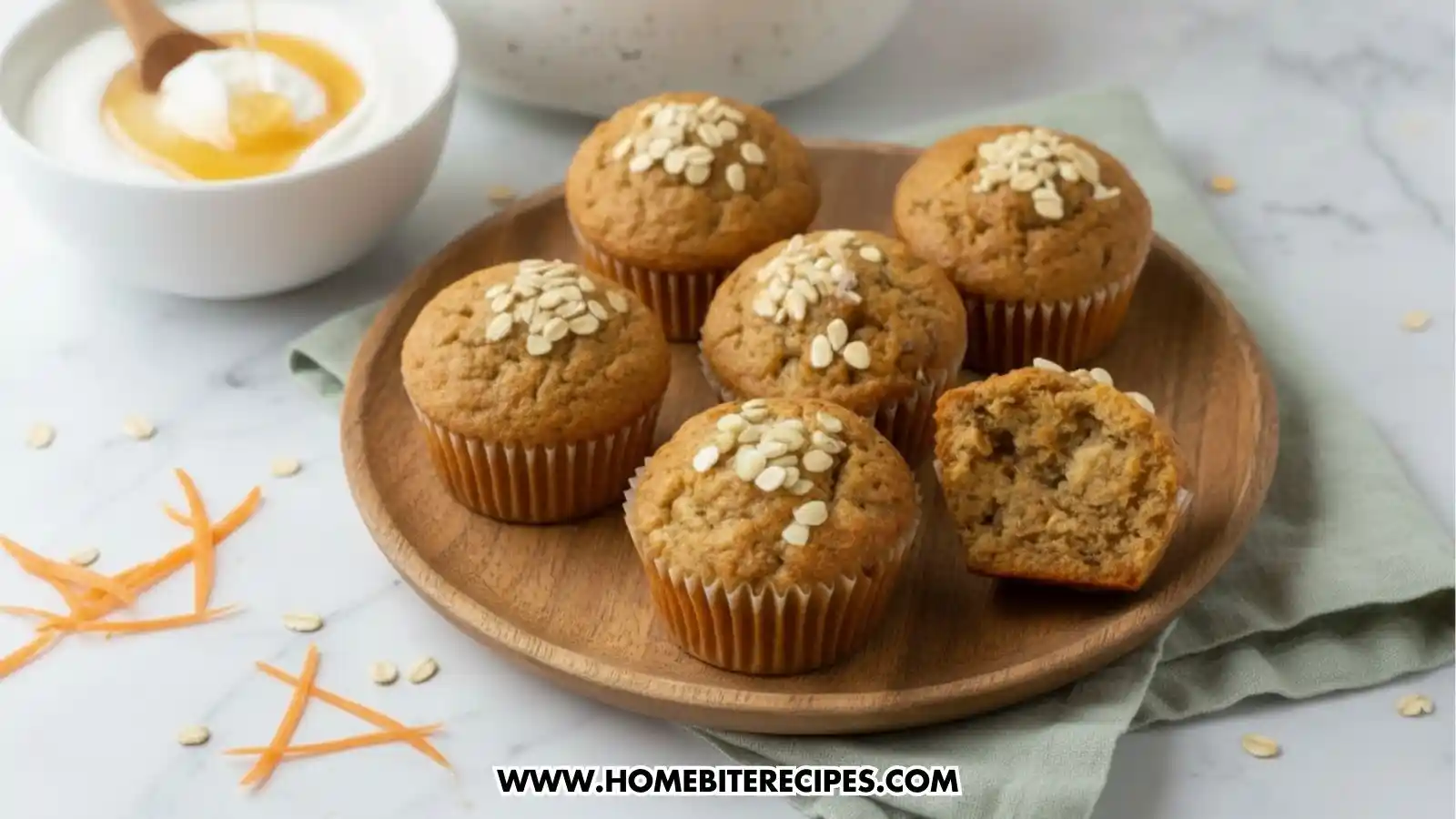 Close-up photo of freshly baked healthy carrot and oat muffins topped with oats, arranged on a white surface with shredded carrots beside them. One muffin is cut open to show the moist, tender texture inside. They're served with a creamy bowl of yogurt sweetened with a drizzle of honey