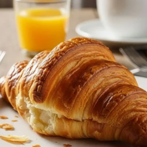 Golden, flaky French croissant served on a white plate with a glass of orange juice and a cup of coffee in the background, creating a classic Parisian breakfast scene.