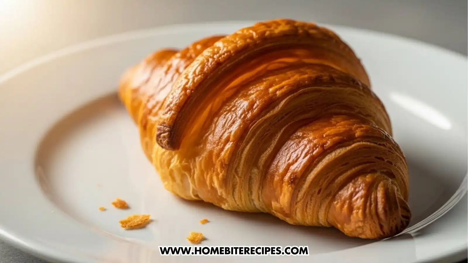 Golden, flaky French croissant served on a white plate with a glass of orange juice and a cup of coffee in the background, creating a classic Parisian breakfast scene.