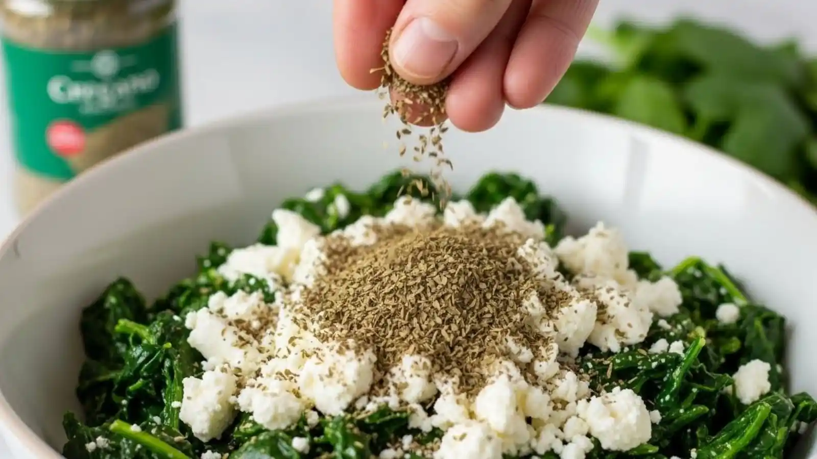 A hand sprinkling dried Mediterranean herbs over a bowl of fresh spinach and crumbled feta cheese, showing a key Mediterranean Spinach and Feta Cheese Crisps step.