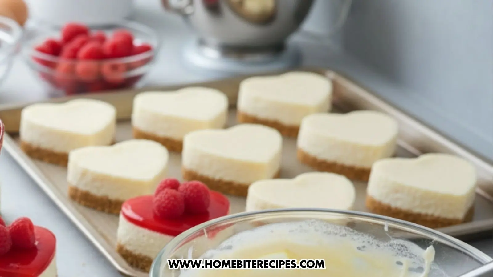 A baking sheet holds several plain heart-shaped cheesecakes on graham cracker crusts, awaiting their toppings. In the foreground, one finished cheesecake with red glaze and raspberries is visible next to a bowl of creamy white batter and a bowl of fresh berries.