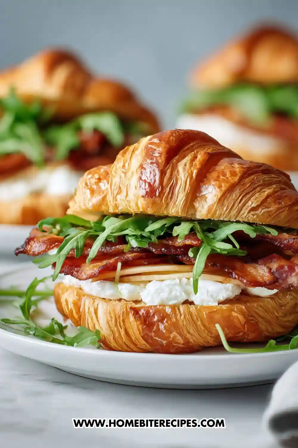 Savory croissant sandwich filled with crispy bacon, creamy ricotta cheese, and fresh arugula on a white plate.