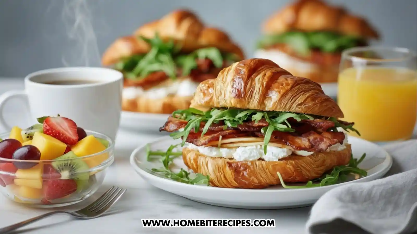 A buttery, golden-brown croissant sandwich filled with layers of crispy bacon, creamy white cheese, and fresh arugula on a white plate. The brunch spread includes a small bowl of mixed fresh fruit (kiwi, strawberry, mango, grape), a white mug of steaming coffee, and a glass of orange juice on a marble tabletop.