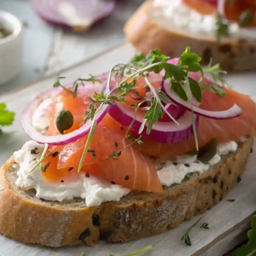 Smoked salmon toast topped with cream cheese, red onion slices, capers, fresh herbs, and black sesame seeds on rustic bread.