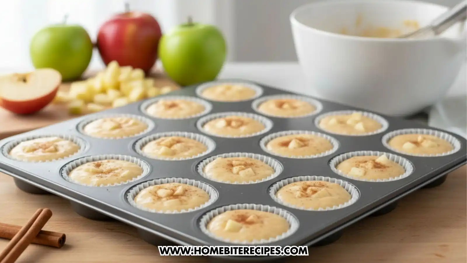 A 12-cup muffin tin filled with apple cinnamon muffin batter ready for the oven, surrounded by fresh apples and cinnamon sticks.