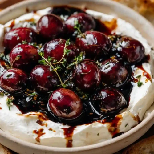 Creamy whipped brie dip in a ceramic bowl topped with glossy roasted balsamic grapes, drizzled with balsamic reduction and fresh thyme, surrounded by toasted baguette slices and rustic crackers